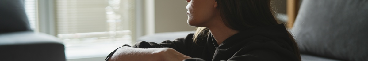 A woman sits with arms folded around her knees at the foot of a bed.